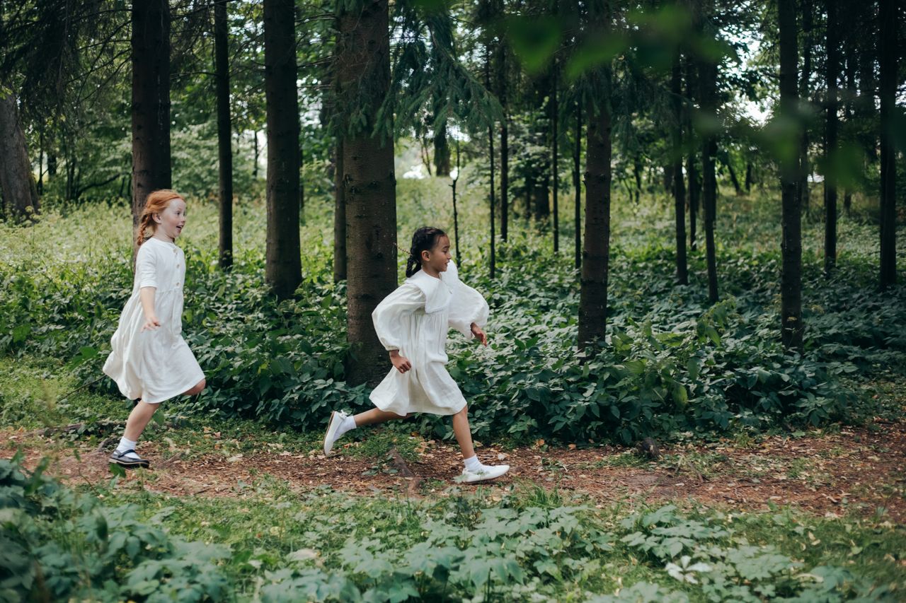 Young girls in white dresses joyfully running through a lush green forest path.
